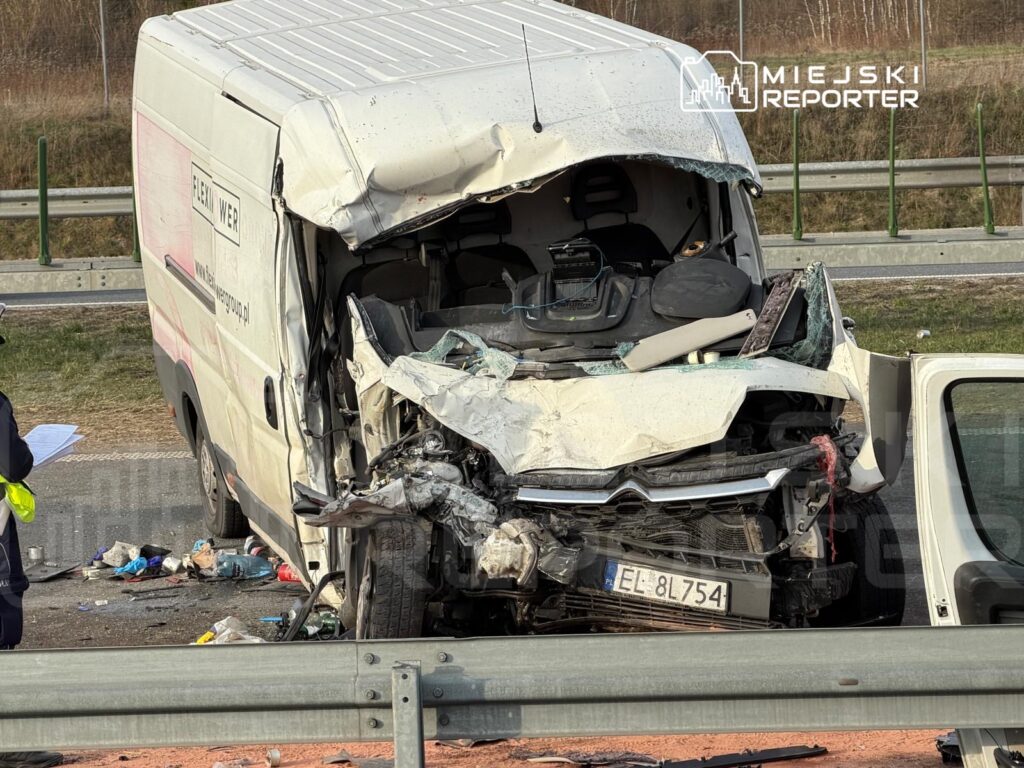 Zniszczony furgon z wgniecionym przodem stoi na poboczu drogi, obok niego pracuje ratownik z notatnikiem.