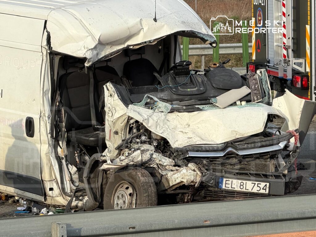 Zniszczony furgon z poważnymi uszkodzeniami przodu, stojący na poboczu drogi, otoczony odpadkami.