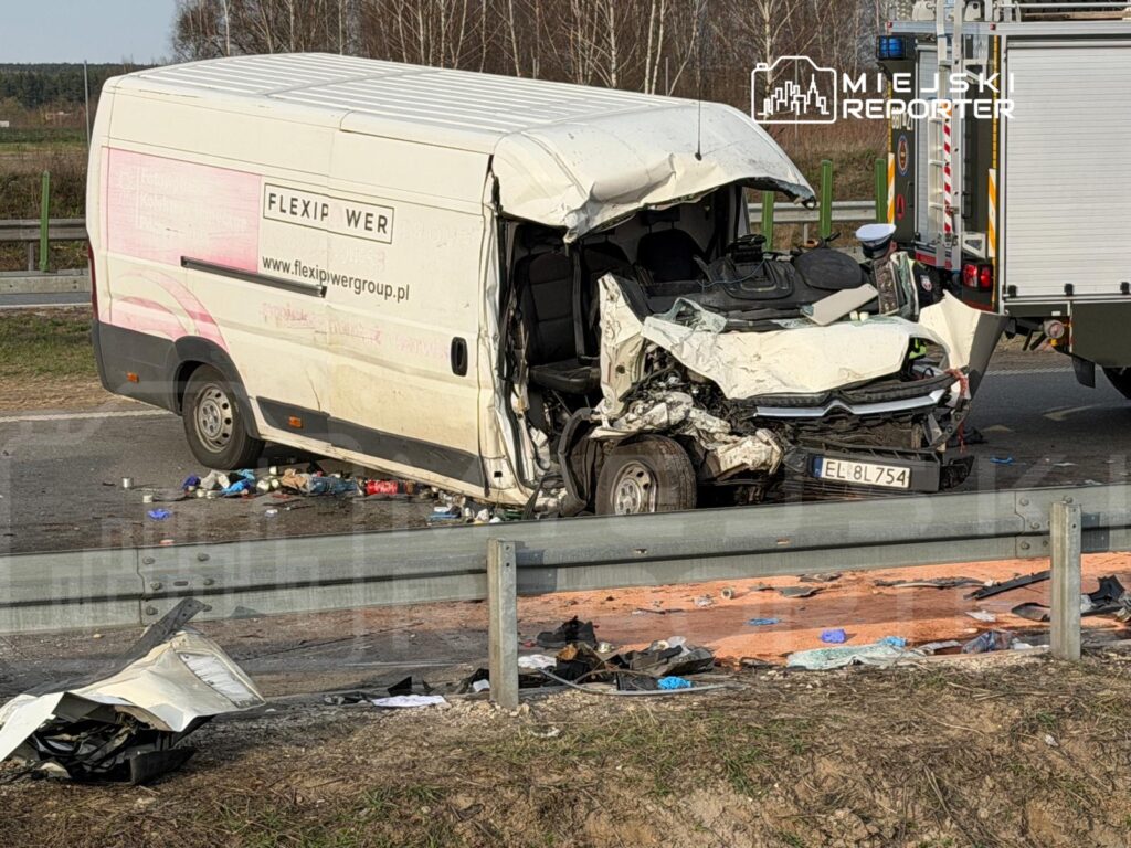 Zniszczony furgon z wgniecionym przodem leży na poboczu drogi, wokół porozrzucane odpady oraz uszkodzone elementy pojazdu.