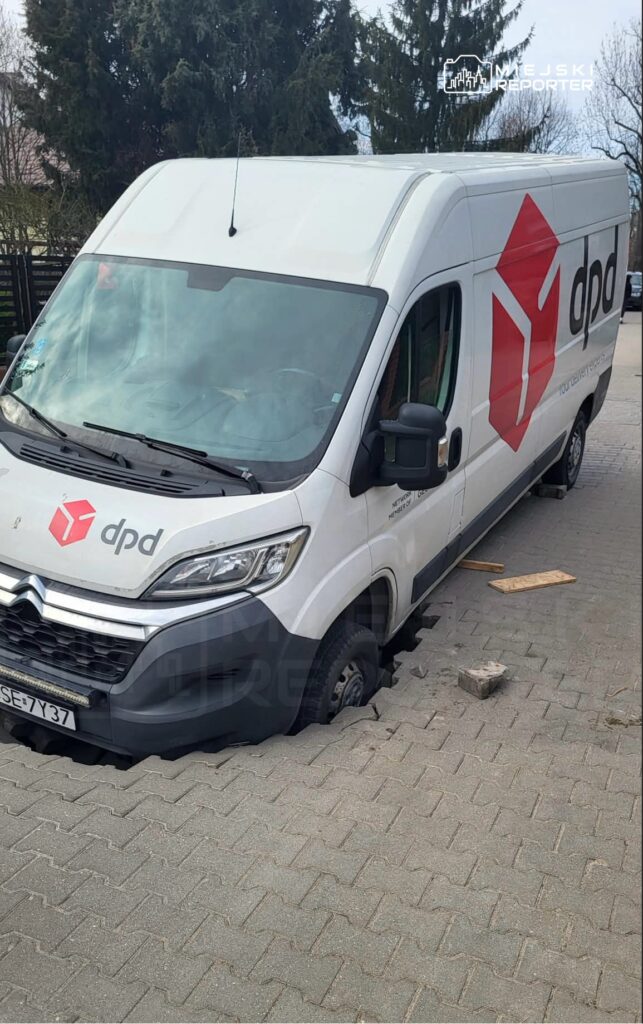 Furgonetka kurierska utknęła w zapadniętym chodniku, z widocznymi uszkodzeniami kół na wybrukowanej nawierzchni.