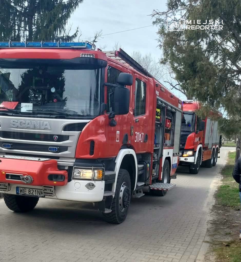 Czerwony wóz strażacki marki Scania stoi na chodniku, obok niego drugi wóz strażacki, w tle drzewa.