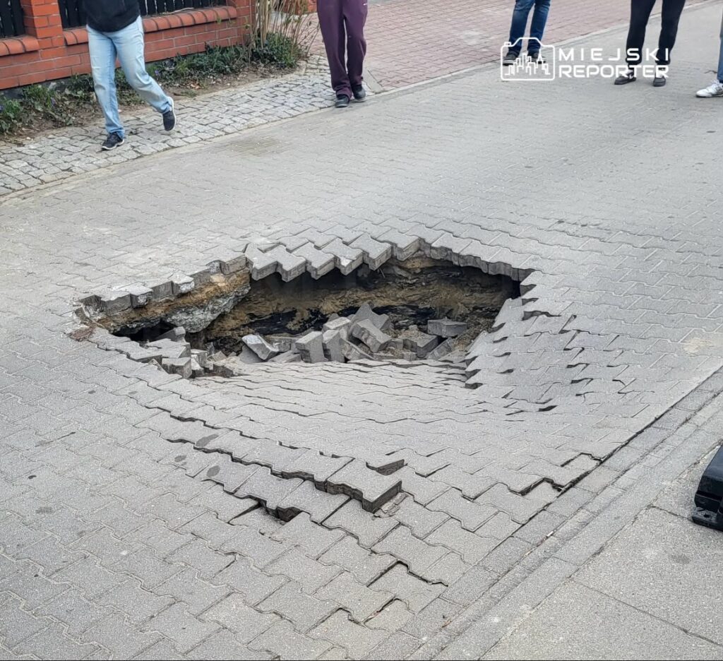 Grupa osób spaceruje obok dużego zapadniętego fragmentu chodnika, odsłaniającego ziemię i zniszczone kostki brukowe.