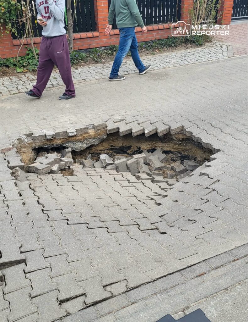 Dwie osoby idą obok dużego zapadniętego fragmentu chodnika, odsłaniającego podziemne warstwy.