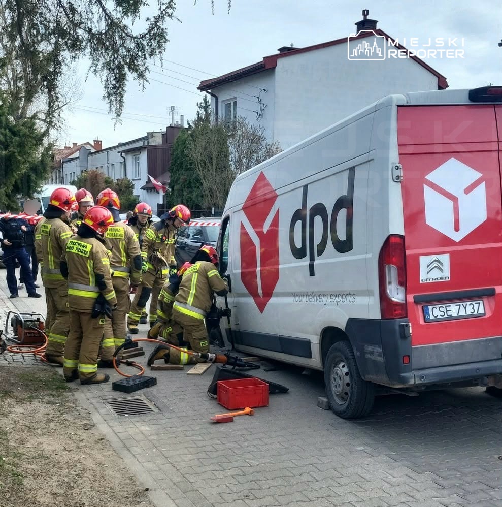 Grupa strażaków w specjalnych strojach pracuje nad otwarciem drzwi furgonetki DPD na miejskiej ulicy.