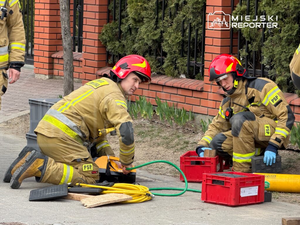Dwaj strażacy w hełmach i mundurach roboczych przygotowują sprzęt na chodniku obok ogrodzenia.