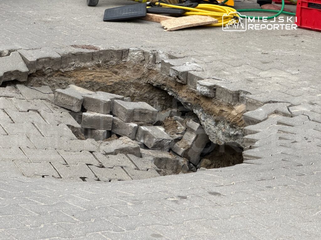 Otwór w nawierzchni z kostki brukowej, odsłaniający podłoże, z uszkodzonymi fragmentami otaczających kostek.