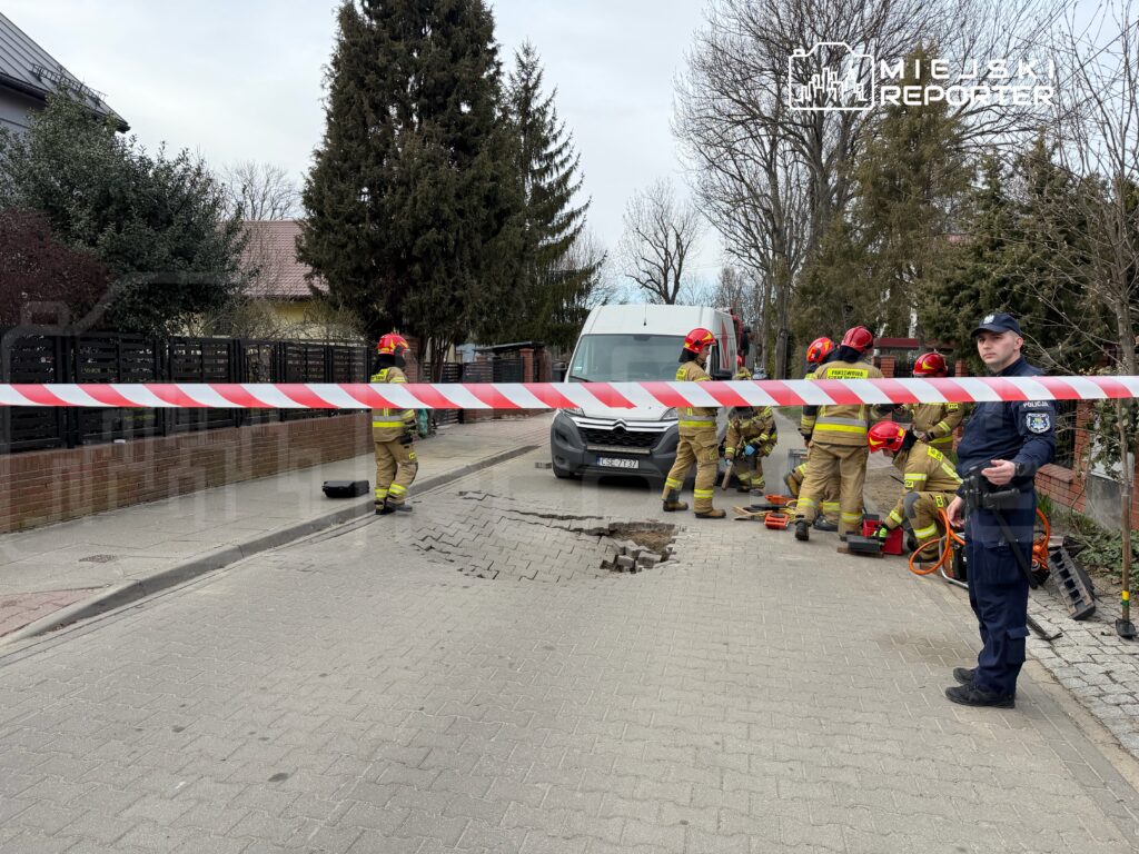 Strażacy w kombinezonach pracują nad usunięciem usterki w jezdni, podczas gdy policjant zabezpiecza teren.