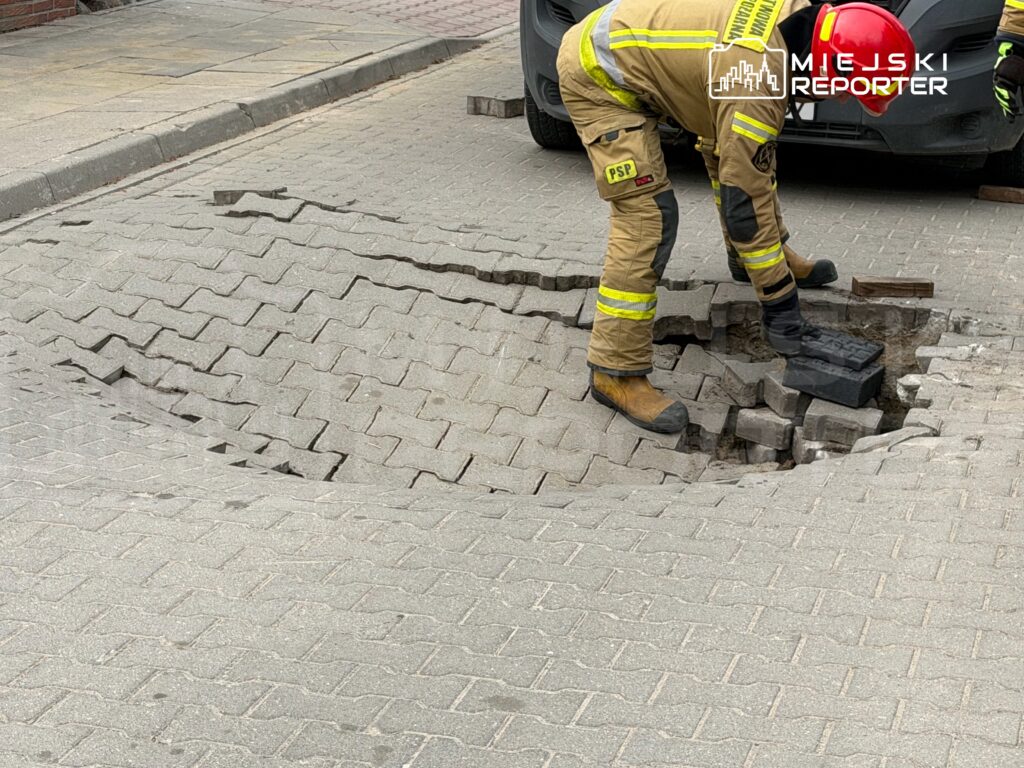 Strażak naprawia uszkodzoną nawierzchnię z kostki brukowej na ulicy, obok zaparkowanego samochodu.