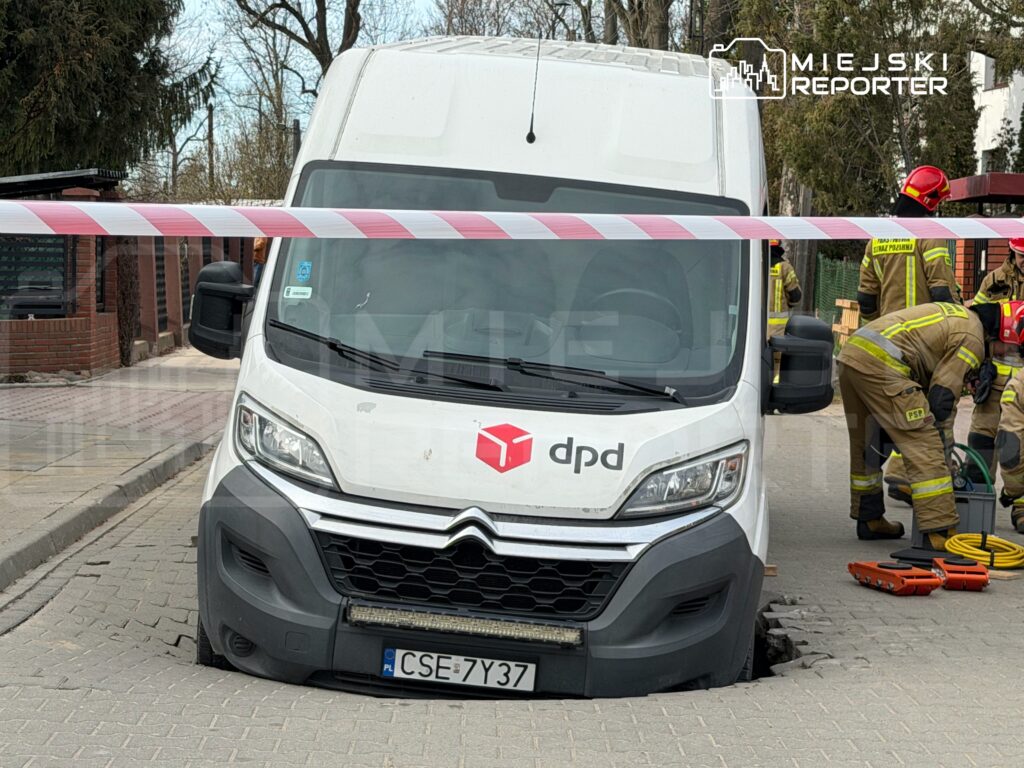 Samochód dostawczy utknął w dziurze na ulicy, a obok pracują strażacy w żółtych mundurach.