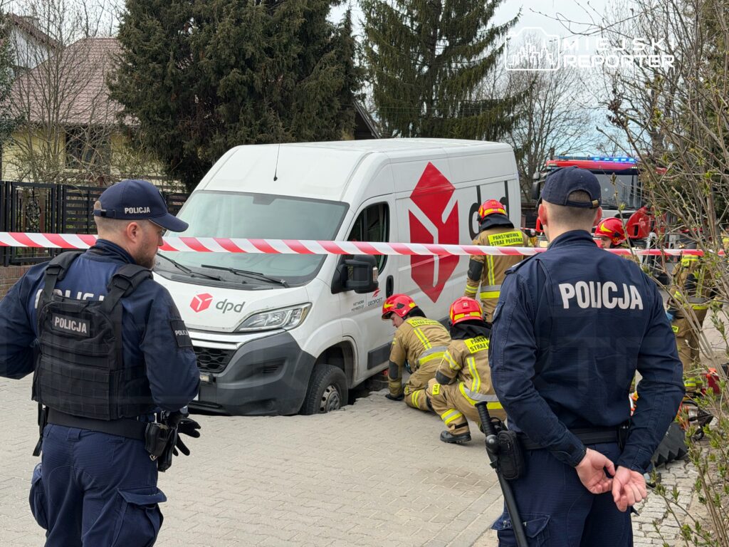 Policjanci obserwują akcję ratunkową, w której strażacy pracują przy samochodzie dostawczym z uszkodzoną oponą.