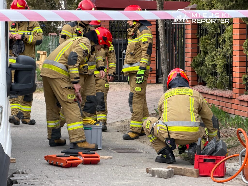Grupa strażaków w mundurach roboczych przygotowuje sprzęt na chodniku przed budynkiem mieszkalnym.
