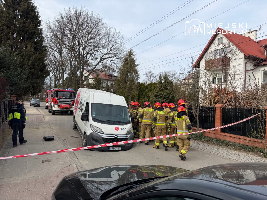 Grupa strażaków w hełmach podchodzi do zaparkowanego vana dostawczego na ulicy, obok wozu strażackiego.
