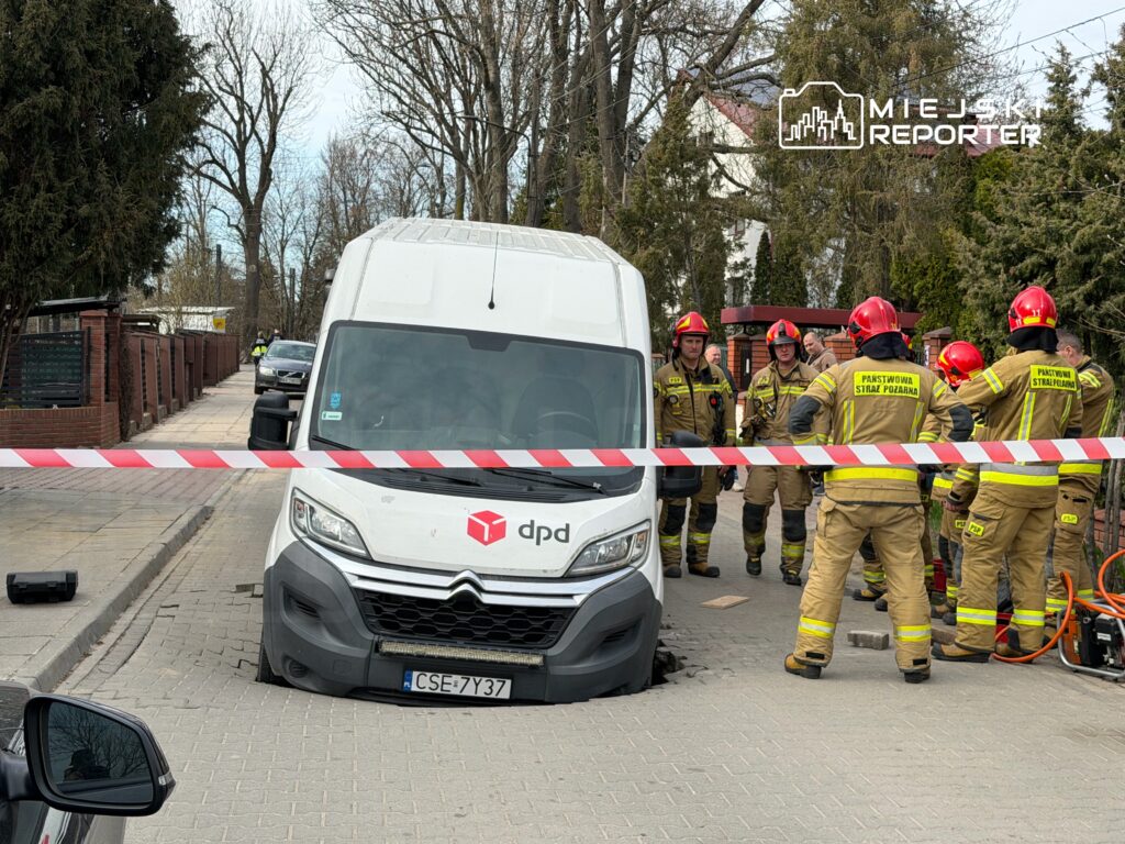Samochód dostawczy utknął w dziurze na ulicy, obok grupy strażaków w hełmach, przygotowujących się do akcji ratunkowej.