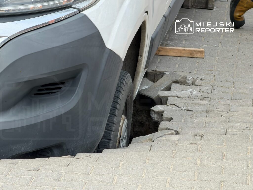 Biały pojazd utknął w uszkodzonej nawierzchni, odsłaniając fragment wykopanej ziemi obok krawężnika.