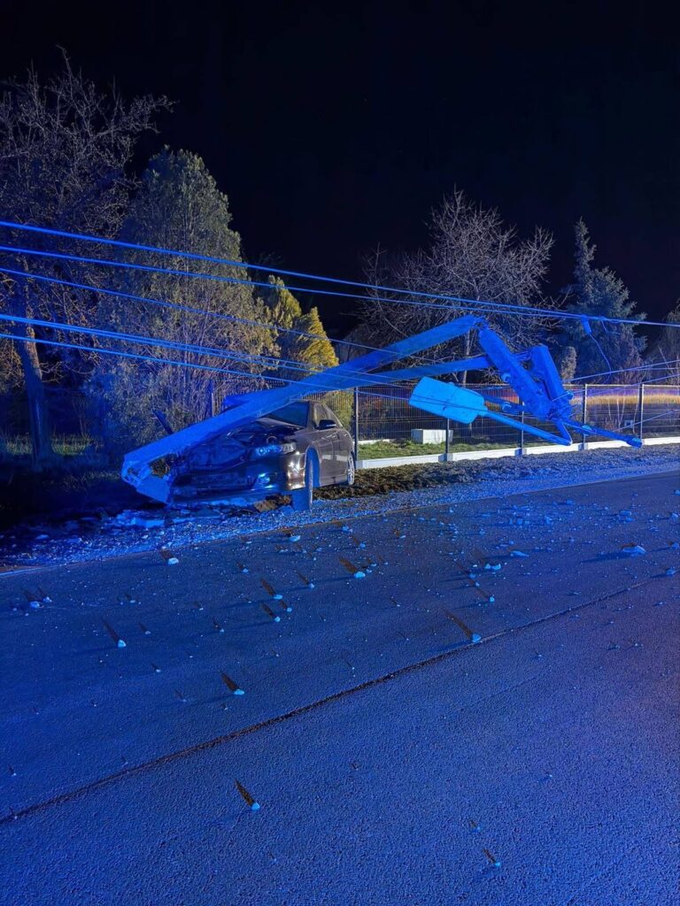 Zniszczony samochód leży na poboczu drogi, a nad nim zwisają uszkodzone kable energetyczne. W tle widoczne drzewa.
