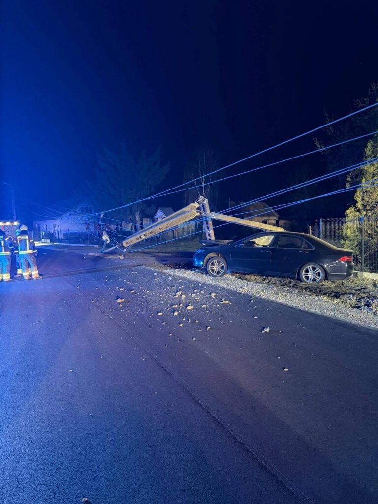 Złamany słup energetyczny leży na drodze obok czarnego samochodu, a strażacy zabezpieczają miejsce zdarzenia w nocy.
