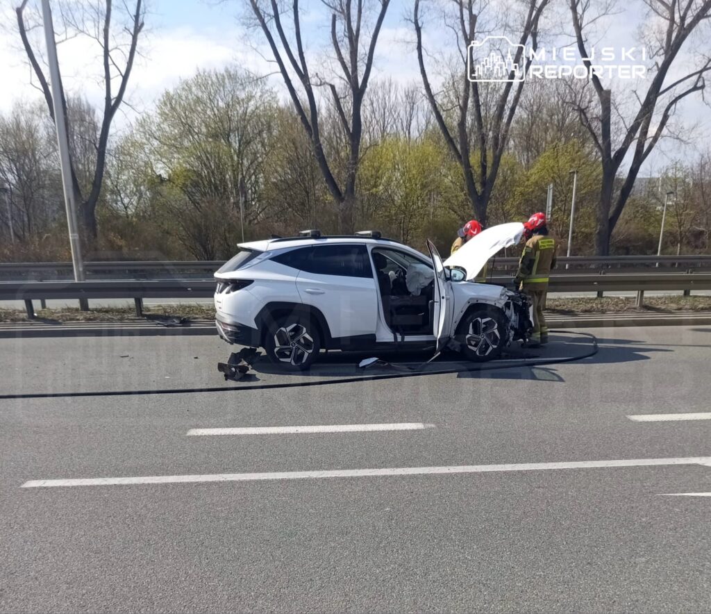 Biały samochód osobowy z uszkodzoną przednią częścią stoi na drodze, obok niego pracują strażacy w hełmach.