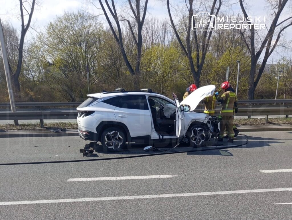 Strażacy pracują przy uszkodzonym samochodzie osobowym na drodze, obok widać odłamki i elementy pojazdu.