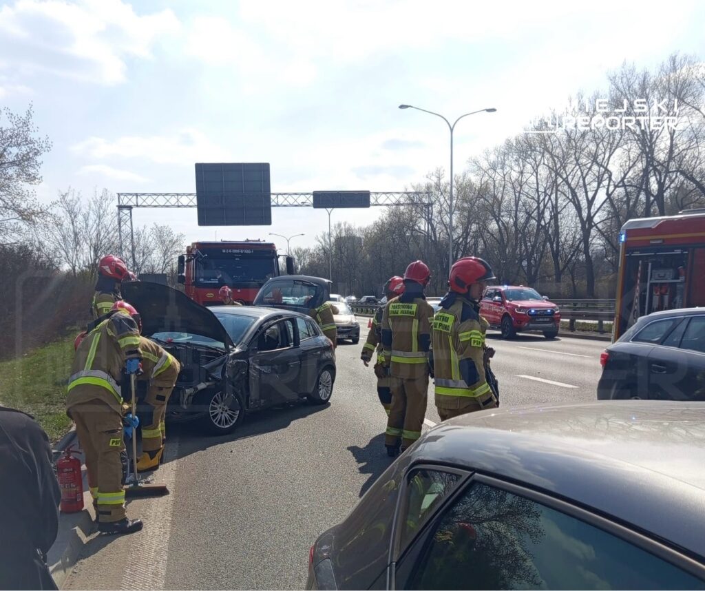 Grupa strażaków w hełmach udziela pomocy przy wypadku drogowym na ruchliwej drodze.