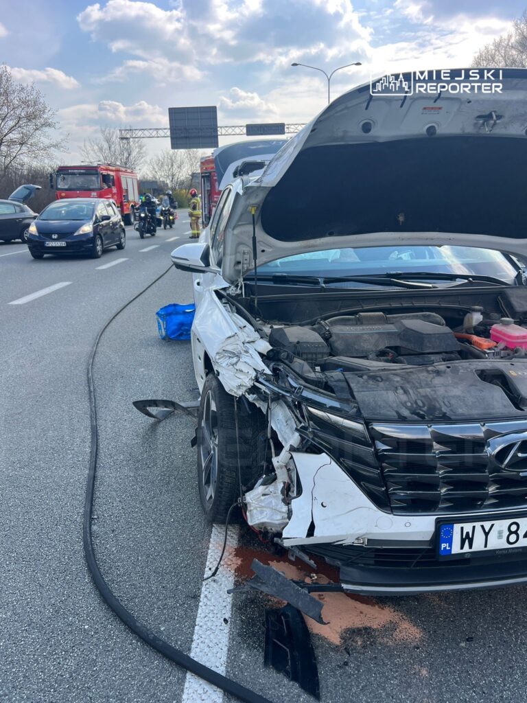 Uszkodzony samochód osobowy z wgniecioną przednią częścią stoi na pasie ruchu, obok interweniują strażacy.