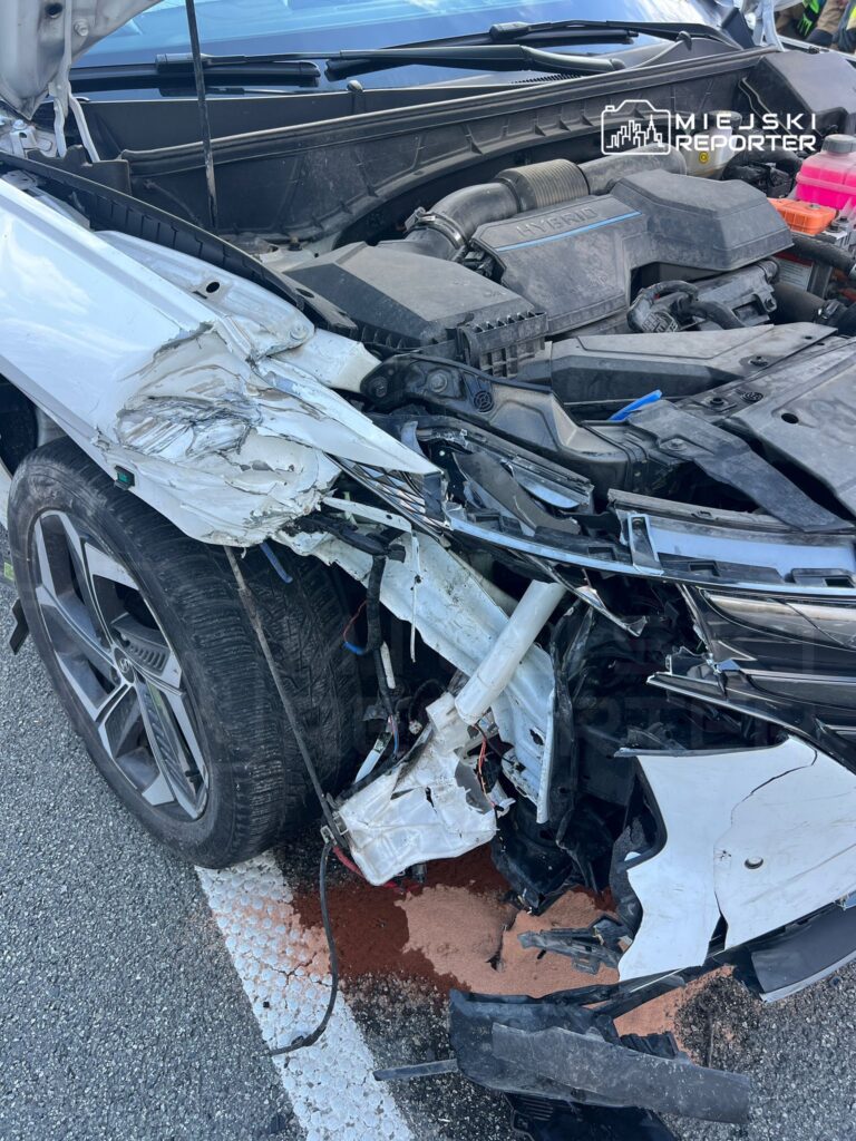 Uszkodzona biała hybryda z wgniecionym przodem, leżąca na drodze, z plamą płynu pod silnikiem.