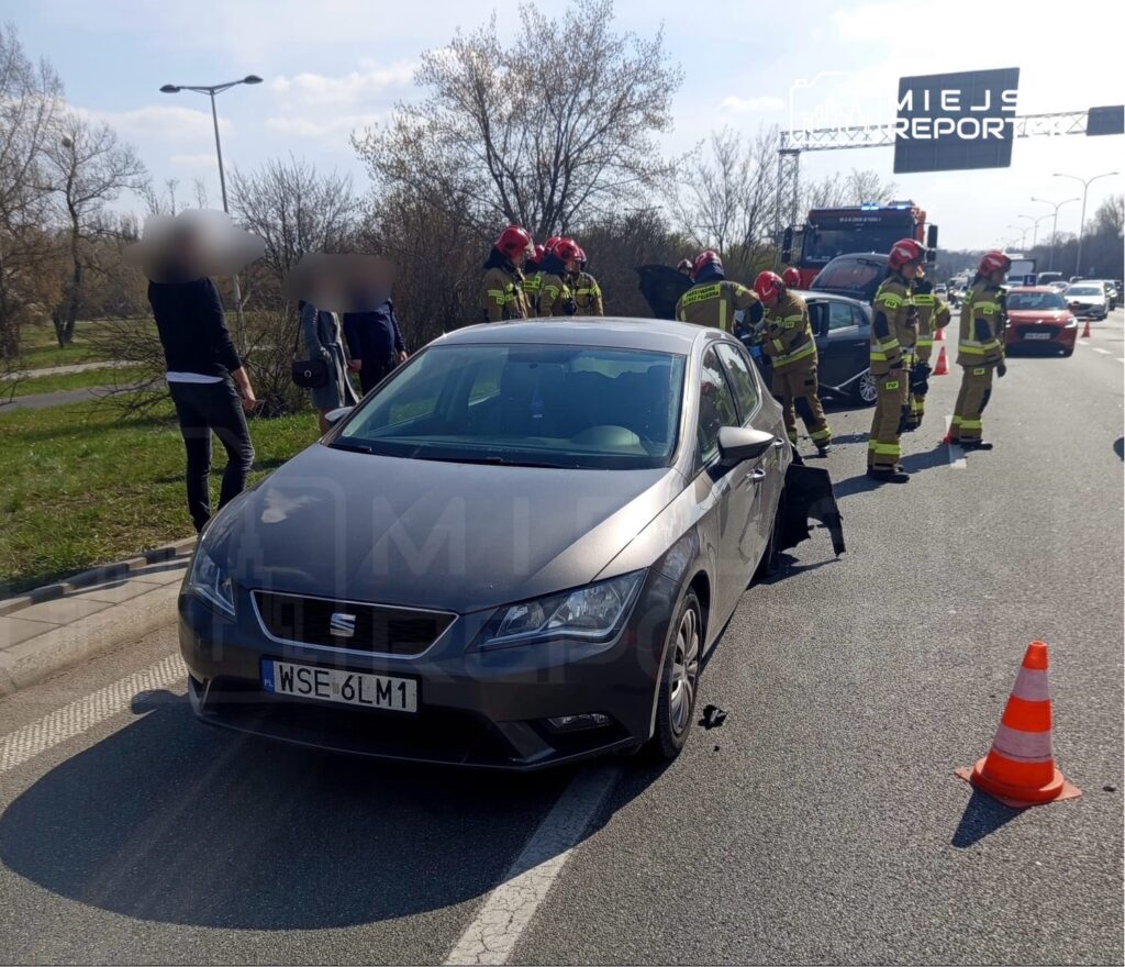 Grupa strażaków w kaskach z czerwonymi hełmami interweniuje przy uszkodzonym samochodzie osobowym na drodze.