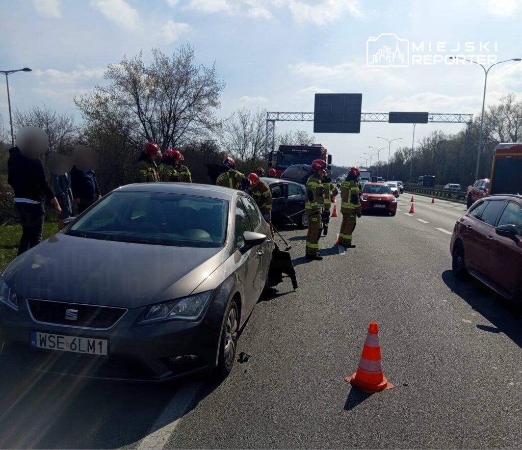 Grupa strażaków w hełmach udziela pomocy poszkodowanym na drodze, obok uszkodzonego samochodu.