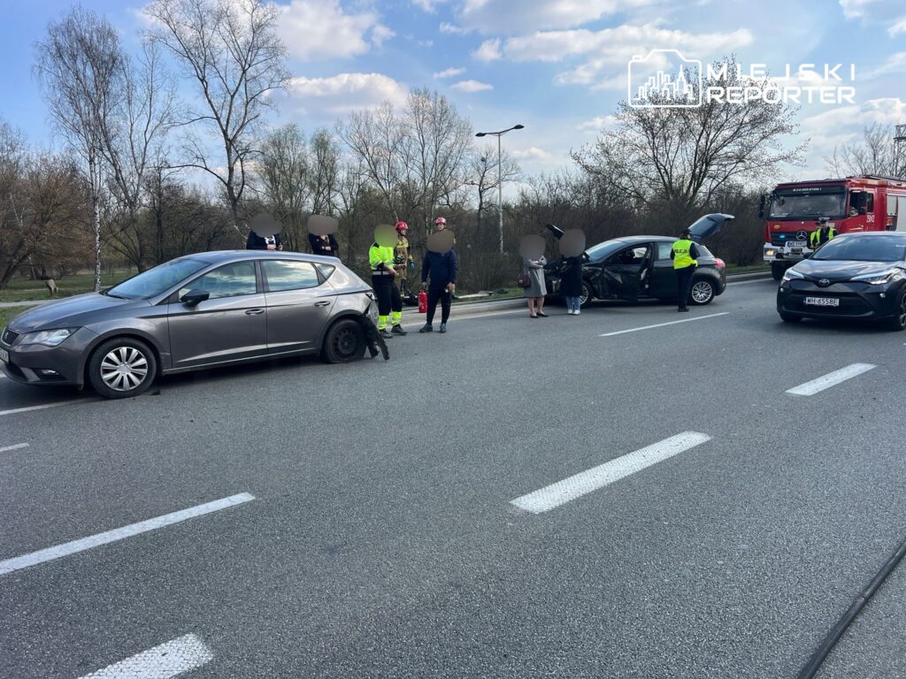 Na drodze stoi uszkodzony srebrny samochód z przebitą oponą, obok grupka osób oraz służby ratunkowe.