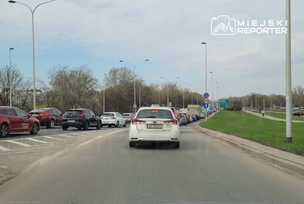 Ruch samochodowy w kierunku centrum, z długim zatorze na drodze, otoczony drzewami i znakami drogowymi.