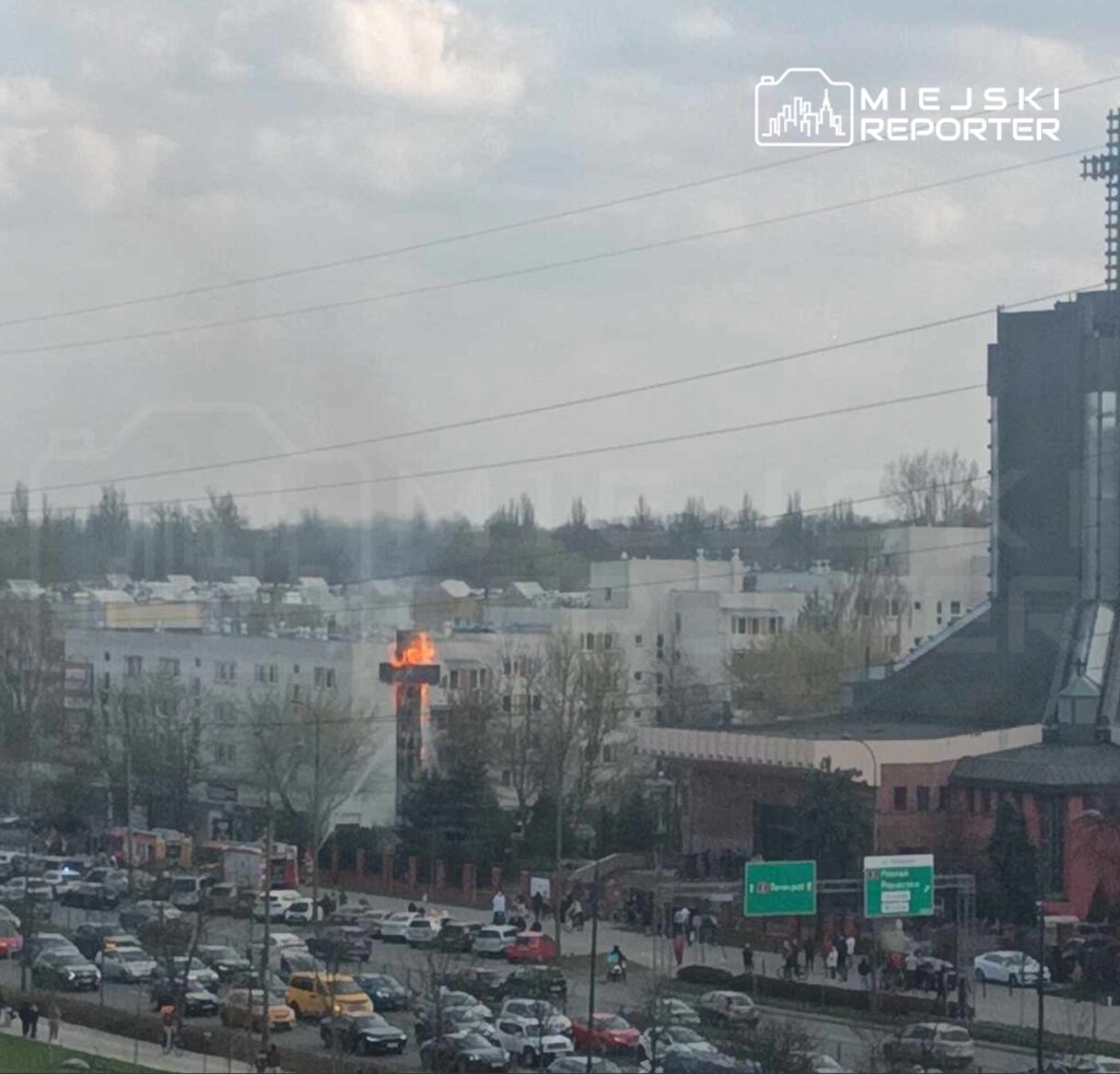 Na dachu budynku widoczny jest ogień, a w tle zgromadzeni ludzie oraz pojazdy na ulicy.