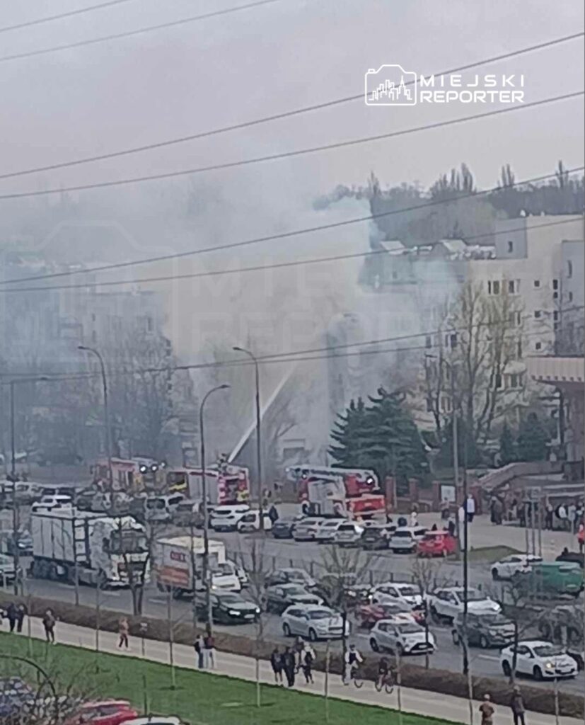W tle widoczny dym unoszący się z miejsca pożaru, obok strażacy używający sprzętu gaśniczego w miejskim otoczeniu.
