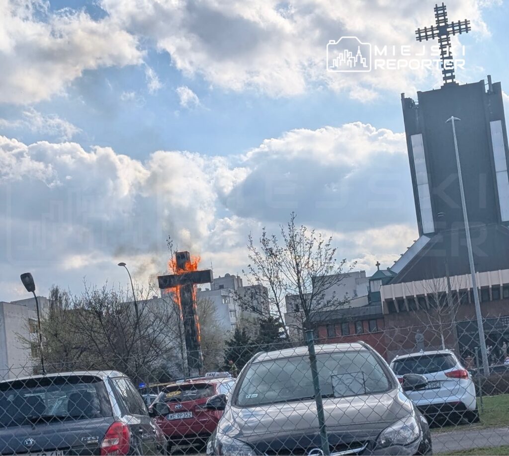 Płonący krzyż na tle chmur, z widokiem na budynki i zaparkowane samochody w pobliżu kościoła.