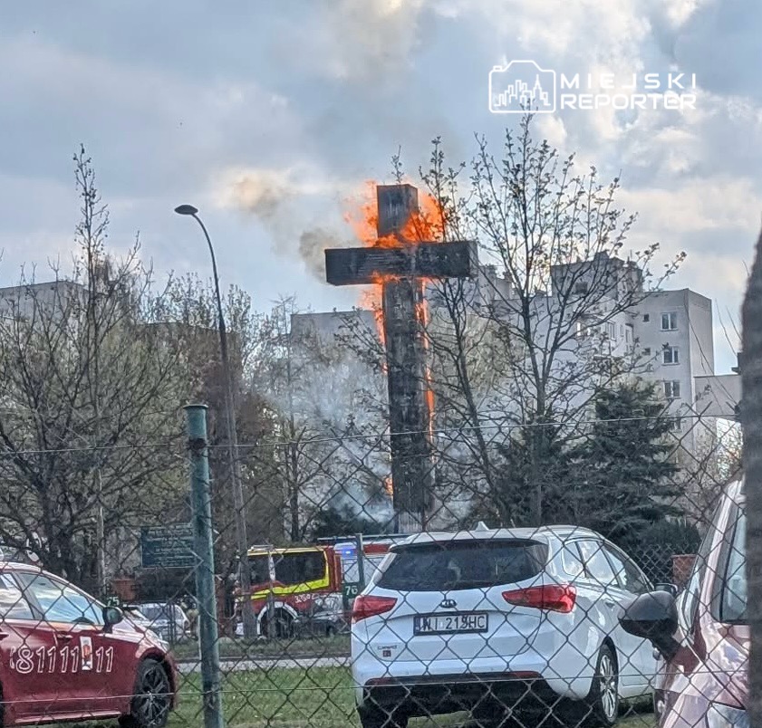 Płonący krzyż w otoczeniu drzew i budynków, w tle widoczny wóz strażacki.