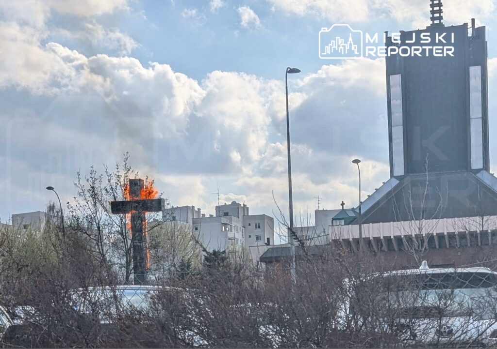 Płonący krzyż wśród budynków mieszkalnych, w tle widoczny kościół oraz chmury na niebie.