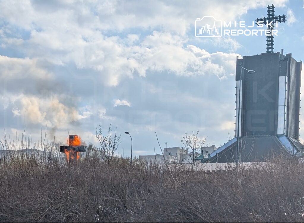 Płonący krzyż na tle nieba z chmurami i nowoczesnym budynkiem w tle, otoczony nagimi krzewami.