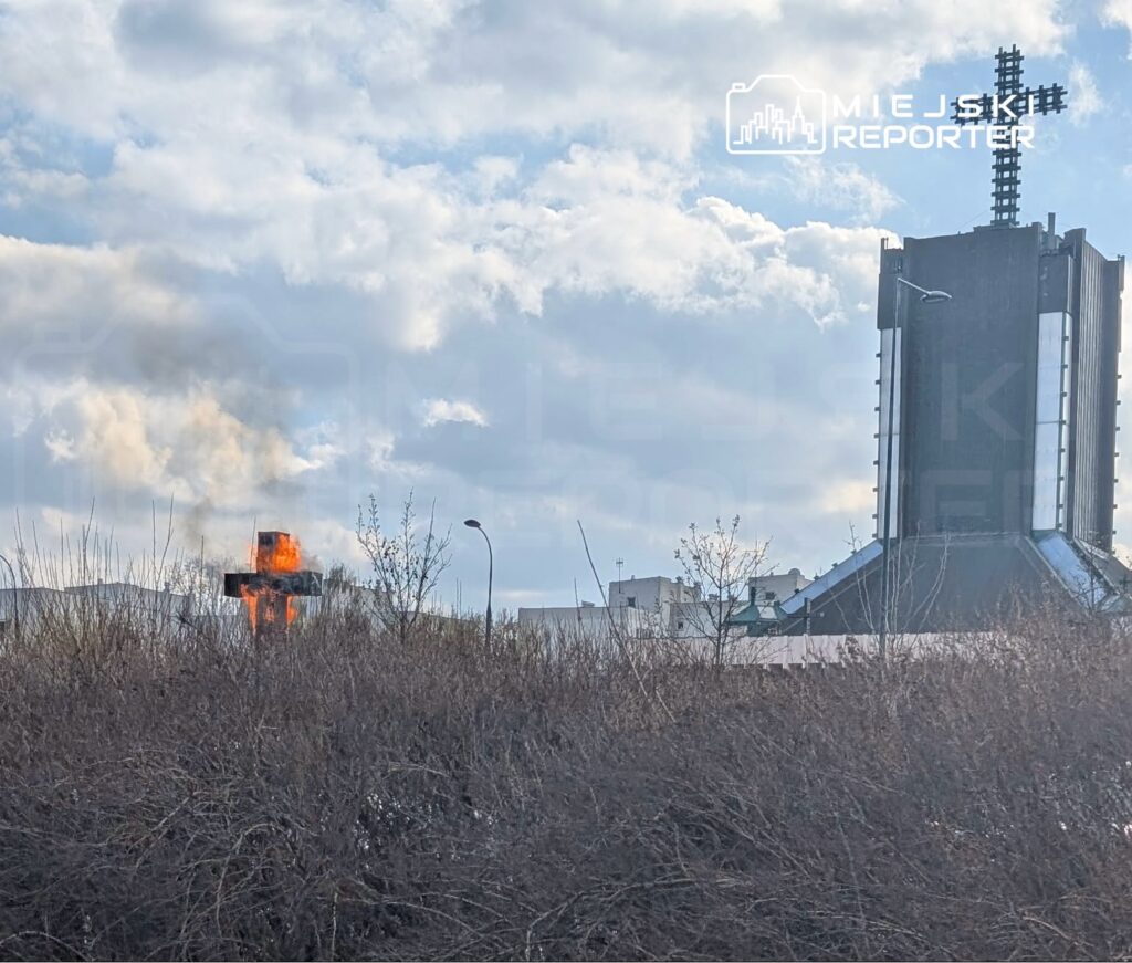 Płonący krzyż na tle nowoczesnego budynku w chmurzystym otoczeniu.