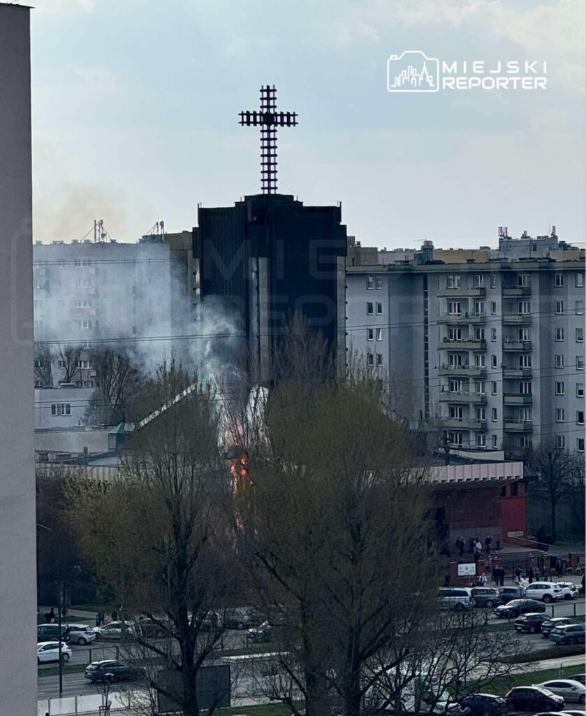 Dym i płomienie wydobywają się z budynku z krzyżem na dachu, w otoczeniu bloków mieszkalnych.