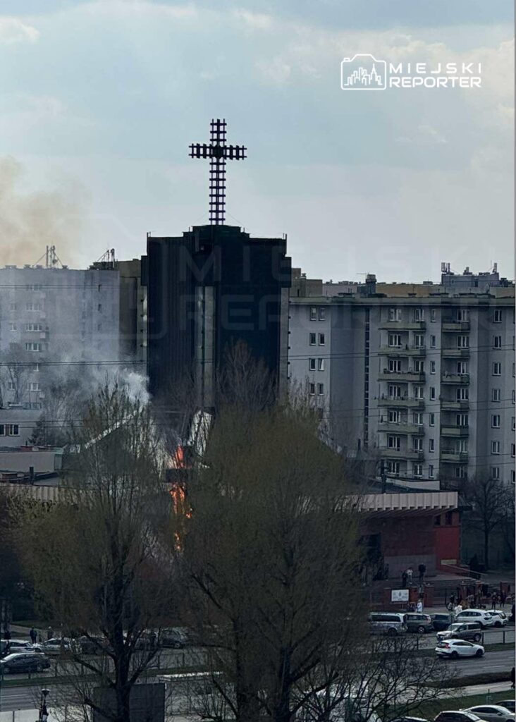 Kościół z dużym krzyżem na dachu, w tle unoszący się dym i płomienie, otoczony blokami mieszkalnymi.