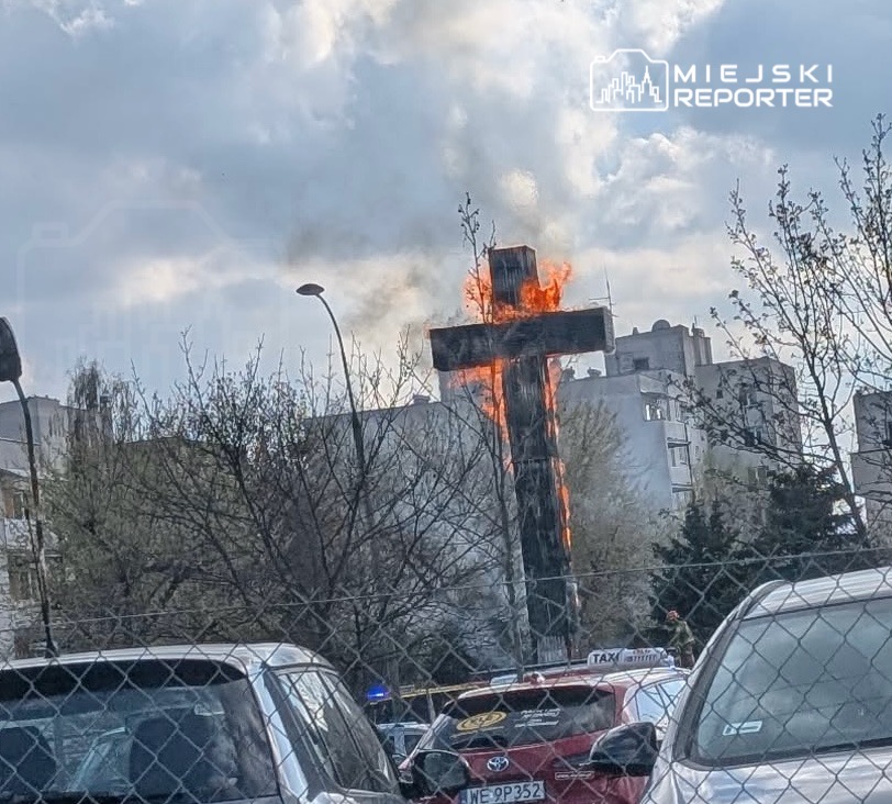 Płonący krzyż otoczony drzewami i samochodami na parkingu, w tle widoczne budynki mieszkalne.
