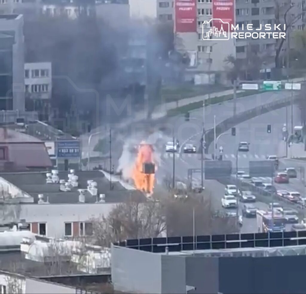 Płonący budynek w miejskim otoczeniu, z dymem unoszącym się w powietrzu, w tle ruchliwa ulica.
