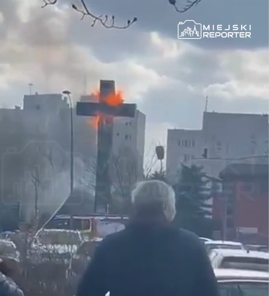 Mężczyzna obserwujący płonący krzyż na tle budynków mieszkalnych w pochmurny dzień.