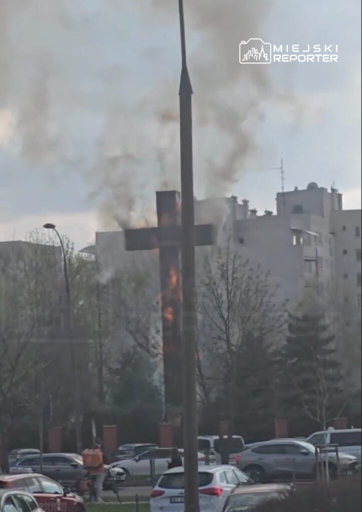 Krzyż płonący na tle miejskiego krajobrazu, z unoszącym się dymem wśród budynków mieszkalnych.