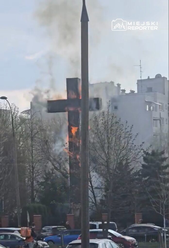 Płonący krzyż na tle budynków, wydobywający się dym w otoczeniu drzew i zaparkowanych samochodów.