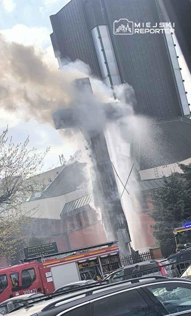 Dym wydobywający się z budynku, w tle widać wóz strażacki i znak informacyjny.