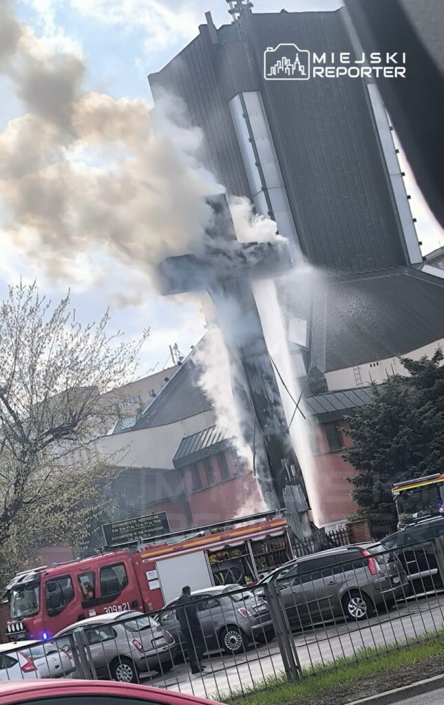 Wysoka wieża z krzyżem wydobywa dym, podczas gdy strażacy gaszą pożar przy budynku kościoła.