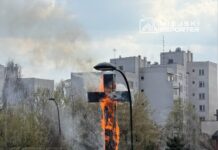Płonący krzyż w tle budynków, z dymem unoszącym się w powietrzu i samochodami zaparkowanymi w pobliżu.