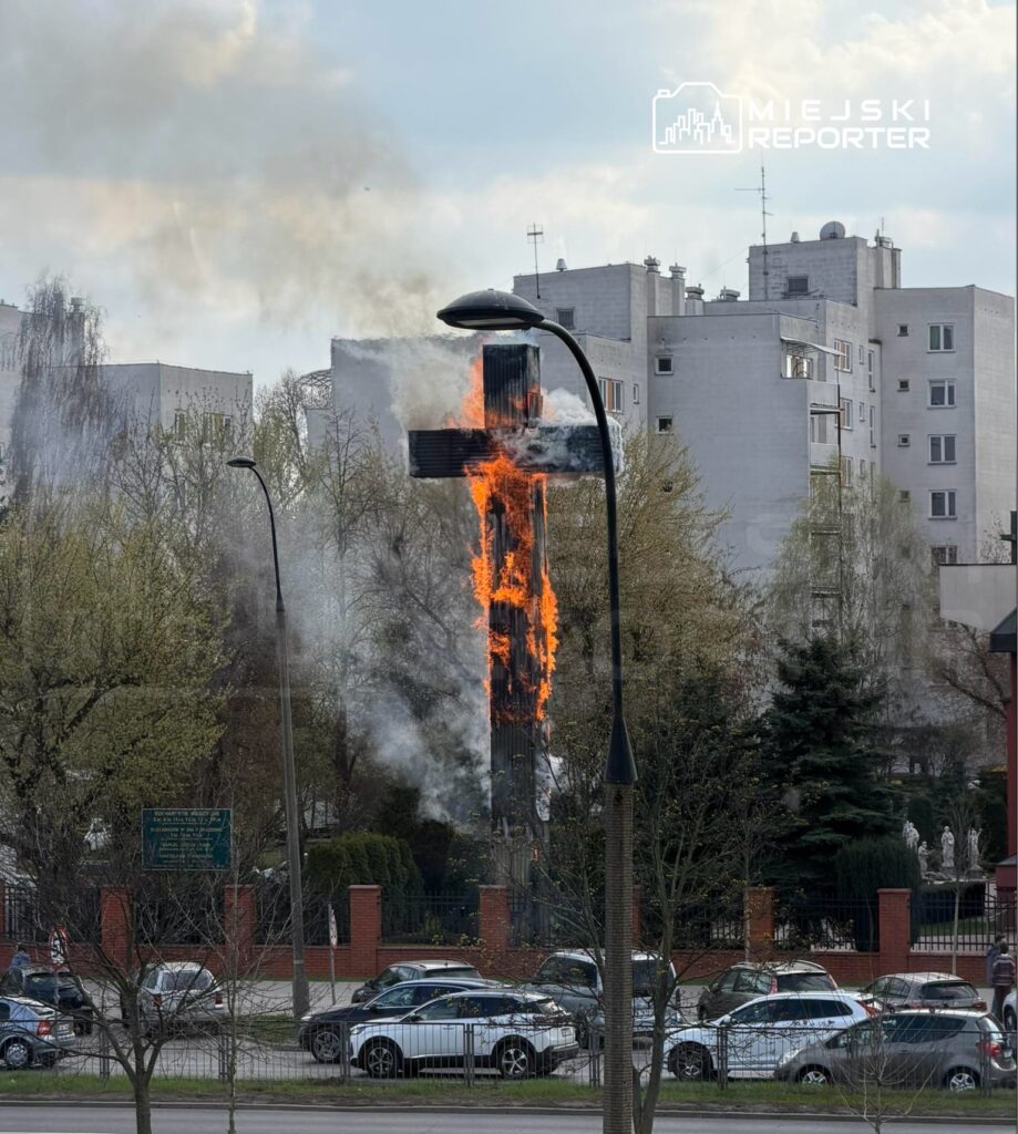Płonący krzyż w tle budynków, z dymem unoszącym się w powietrzu i samochodami zaparkowanymi w pobliżu.