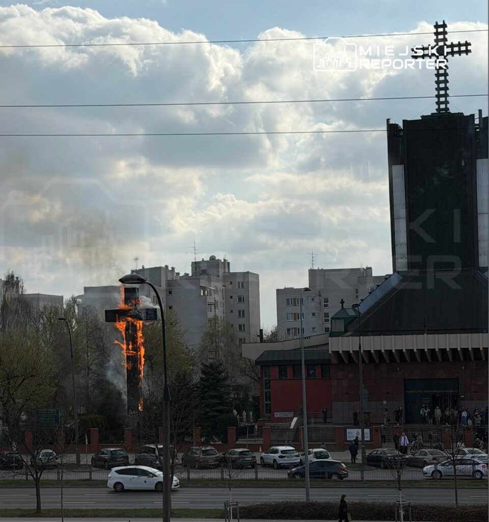 Płonący krzyż na tle budynków mieszkalnych, w pobliżu kościoła, otoczony dymem i płomieniami.