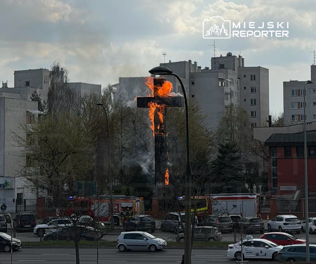 Płonący krzyż z płomieniami i dymem, otoczony budynkami mieszkalnymi, w tle widoczne samochody i wóz strażacki.