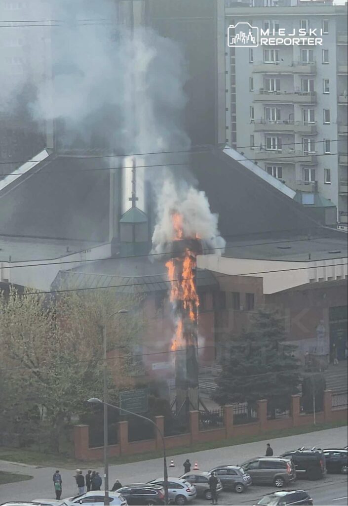 Płonący komin budynku z dymem i ogniem, wokół grupka ludzi obserwujących zdarzenie. W tle nowoczesne bloki mieszkalne.